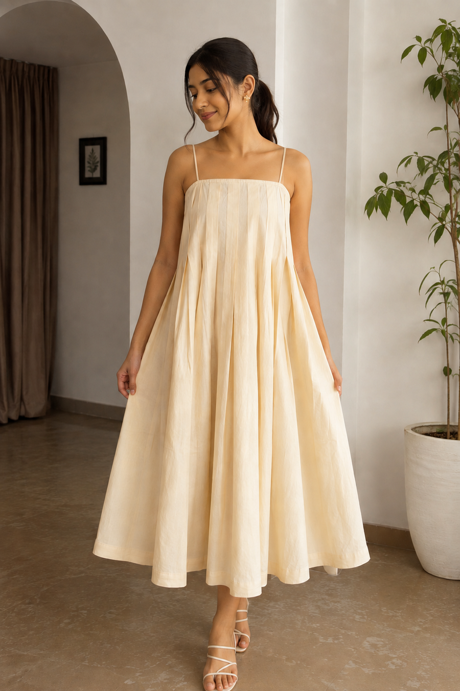 A woman wearing a cream-colored linen midi dress with thin spaghetti straps and a gathered, flowing skirt, standing in a minimalist indoor setting with a potted plant and neutral walls.